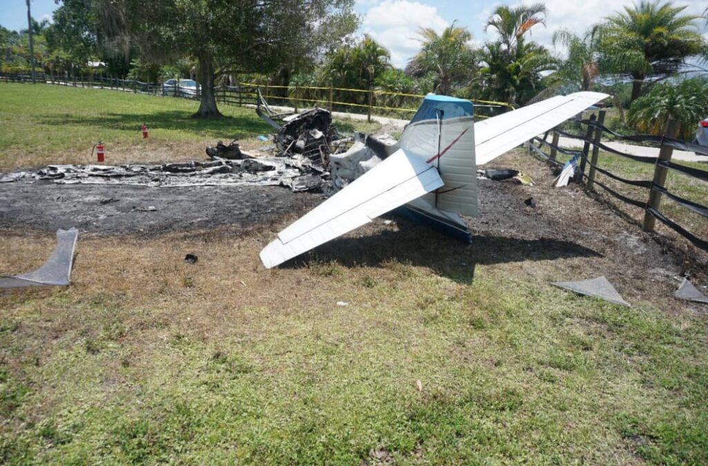 Rear view of Commander 114TC crash site, showing the intact tail section, blackened ground, and scattered fire extinguishers along the wreckage path in a grassy field.